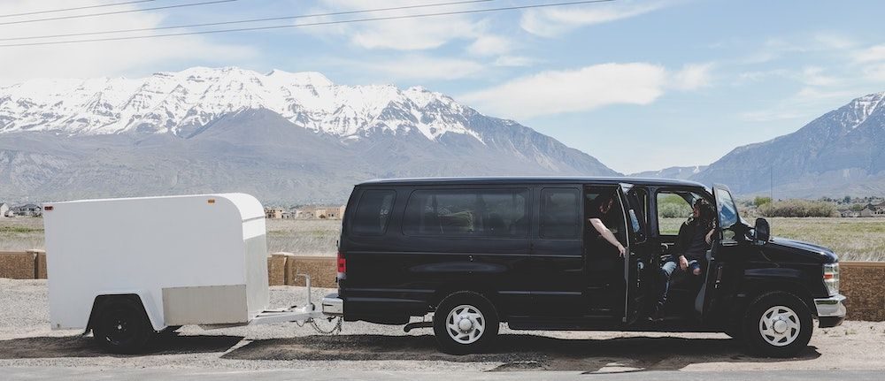Schwarzer Tour-Van mit weißem Equipment-Anhänger vor schneebedeckten Bergen.